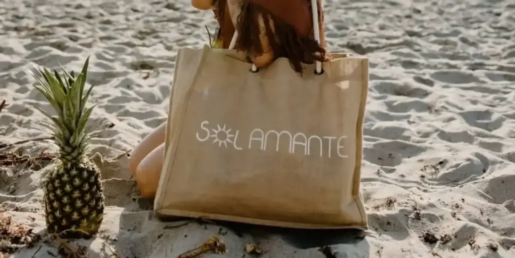 Person at the beach holding a personalized tote bag with "SOLAMANTE" logo, next to a pineapple, showcasing custom bags as trendy promotional items.